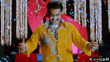 a man in a yellow shirt is giving a thumbs up while blue petals fall on him .
