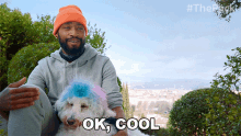 a man is sitting next to a white dog with a blue and pink wig and says " ok cool "