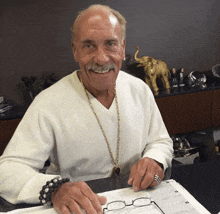 a man wearing a white sweater and a gold chain around his neck sits at a desk