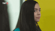 a woman with long black hair is standing in front of a yellow wall with the letters trt1 on the bottom