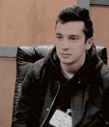 a young man is sitting in a chair wearing a name tag and a jacket .