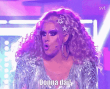 a drag queen is standing on a stage with purple lights behind her and says ' oonna daj ' .