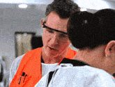 a man wearing an orange vest and safety goggles talks to a woman