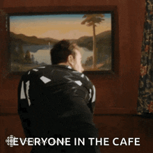a man in a black and white jacket is standing in front of a framed picture and says everyone in the cafe .