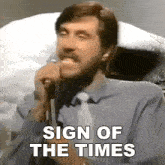 a man singing into a microphone with the words " sign of the times " above him