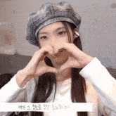 a woman wearing a hat makes a heart shape with her hands in front of her face