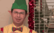 a man wearing a green elf hat and a bow tie is smiling in front of a christmas tree .