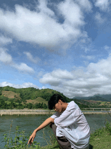 a man is squatting down by a river with a mountain in the background
