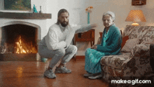 a man is squatting next to an elderly woman sitting on a couch in a living room .