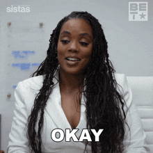 a woman in a white jacket says okay in front of a sign that says sistas