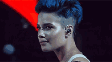 a woman with blue hair and a shaved head is wearing earbuds and a white tank top .
