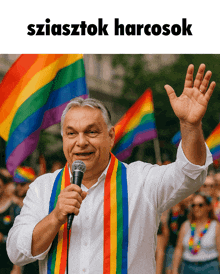 a man with a rainbow scarf around his neck holds a microphone in front of a crowd with rainbow flags
