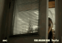 a woman is peeking out of a window with blinds and the words fear walking dead hulu written on the bottom