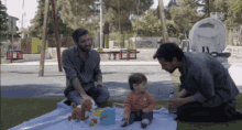 two men are playing with a baby on a blanket in a park and the baby is wearing an orange shirt