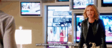 a woman in a leather jacket is standing in front of a wall of televisions and talking .