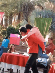 a man in a red jacket is carrying a woman on his back in front of a table