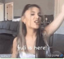 ariana grande is sitting in a living room with her arms outstretched and says yulia is here .