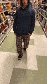 a man wearing plaid pants and a blue hoodie walks down a grocery store aisle