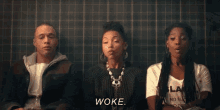 a group of people are sitting next to each other in front of a tiled wall and one of the girls is saying woke .