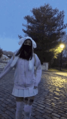 a girl wearing a mask and bunny ears is standing on a cobblestone street .