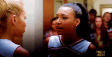 two cheerleaders are standing next to each other in a hallway and talking to each other .