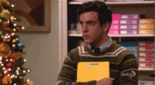 a man in a sweater and tie is holding a clipboard with a bow on it in front of a christmas tree .