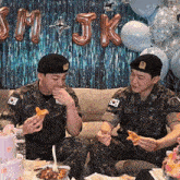 two men in military uniforms sit on a couch eating pizza