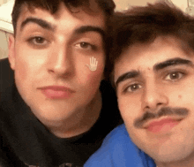 two men with mustaches are posing for a picture together . one of the men has a hand sticker on his face .