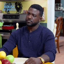 a man in a blue shirt is sitting at a table in a kitchen with fruit .