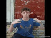 a young man in a blue shirt is sitting in front of a brick wall with his hands outstretched .