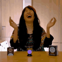 a woman laughs while sitting at a table with a box of magic crystal ball