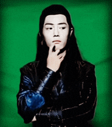 a man with long black hair has his hand on his chin and a green background