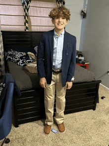a young boy wearing a blue jacket and khaki pants is standing in front of a bed