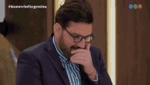 a man in a suit and glasses is covering his mouth while watching a masterchef argentina show