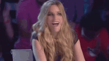 a woman with long blonde hair is laughing and clapping her hands while sitting in a chair .
