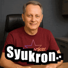a man wearing a red shirt with a star on it sits at a desk with the word syukron written on it