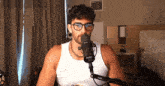 a man wearing glasses and a white tank top is sitting in front of a microphone
