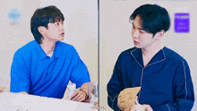 two young men are sitting at a table looking at each other . one of the men is wearing a blue shirt .