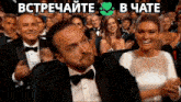 a man in a tuxedo and bow tie is sitting in a crowd of people with the words " встречайте " in green