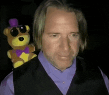 a man in a purple shirt and vest is holding a teddy bear in his pocket .