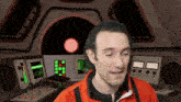 a man in an orange jacket stands in front of a control panel with a red star in the back