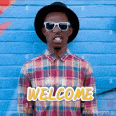 a man wearing sunglasses and a plaid shirt is standing in front of a blue brick wall and says welcome