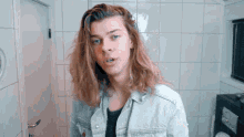 a young man with long blonde hair is standing in front of a white tiled wall