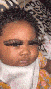 a baby with long eyelashes is wearing a bib and making a funny face .
