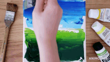 a person is painting on a piece of paper with a brush and a tube of acrylic paint in the background