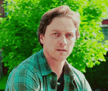 a man in a blue plaid shirt is sitting in front of a tree and looking at the camera .