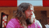 a man with long hair and a beard is wearing a pink shirt and eating a sandwich