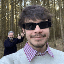 a man wearing sunglasses and a plaid shirt is giving a thumbs up in the woods
