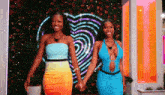 two women are standing next to each other holding hands in front of a heart shaped neon sign .