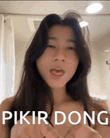 a woman with long hair is making a funny face in a bathroom with the words pikir dong written on her chest .
